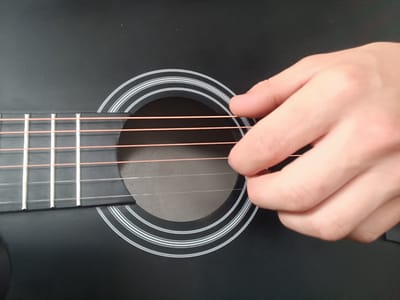A close-up, high-angle shot shows a person's hand playing an acoustic guitar. The guitar is black with six light-colored strings and decorative white rings around the soundhole. The hand is positioned over the strings, with the index finger and thumb visible. The background is a dark, textured surface, possibly the body of the guitar. The lighting is even, highlighting the textures of the guitar and skin. The image is devoid of any context related to the city of Alamüdün, Kyrgyzstan, or any specific urban environment.