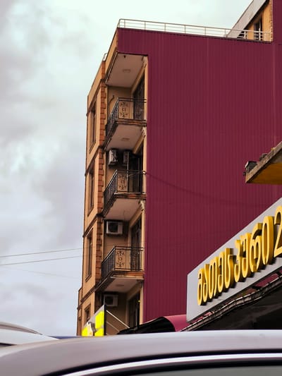 The image depicts the side of a tall, narrow building with multiple balconies, likely an apartment building or a mixed-use structure, in Charnali, Georgia. The building has a facade of light brown brickwork and is topped with a dark red corrugated metal section. Several balconies with ornate black metal railings are visible. Air conditioning units are mounted on the exterior walls beneath some of the balconies. In the foreground, the shiny, curved surface of a vehicle's roof is prominent, partially obscuring the lower portion of the building and a yellow sign with Georgian script. The sky above is overcast with clouds.