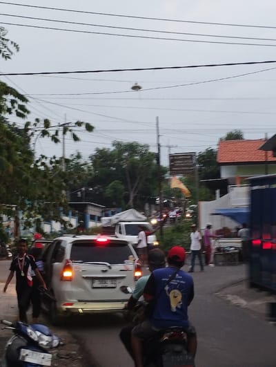 The image depicts a busy street scene in Kecamatan Panji, Indonesia, under an overcast sky, suggesting it is either late afternoon, early evening, or a cloudy day.
In the immediate foreground, the partial view of a dark motorcycle is visible on the left. Two men are riding another motorcycle, seen from behind, in the center-right. The passenger wears a blue t-shirt with a graphic of a lightbulb and an exclamation mark, along with some unreadable text below it. The driver wears a red cap.
To the left of the motorcycle, a white multi-purpose vehicle (MPV), likely a Toyota Avanza, is prominently featured with its brake lights illuminated and a turn signal blinking. Its license plate reads "P 1852 DZ". A man in a black and red shirt, adorned with medals around his neck, stands near the open door of the white car, looking towards the camera.
Further in the background, more people are visible standing or moving, and various buildings with red tiled roofs and blue or white walls line the street. Dense green foliage from trees is present on the left and in the distance. Utility poles and numerous power lines crisscross the gray sky. A small, disc-shaped object, possibly a street light or speaker, hangs suspended from these wires. A yellow or orange flag is visible in the mid-ground, hanging from a pole. On the far right, a section of a blue truck or large vehicle is partially visible. The scene captures a moment of street activity, potentially with some traffic congestion.
