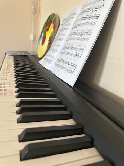 The image depicts a close-up, eye-level view of a digital piano keyboard. The keys are predominantly white, with black keys interspersed. The keyboard is angled diagonally from the bottom left to the top right of the frame. On the left side of the keyboard, the white keys have colored stickers with musical notation and Russian text.
Positioned on the keyboard, to the left of the open sheet music, is a round, wooden object with a colorful painting on it. The painting appears to be abstract, with splashes of yellow, red, and green.
Resting on the right side of the keyboard is an open book of sheet music, displaying musical notes and symbols. The book is angled upwards, leaning against a plain, light-colored wall. The lighting suggests an indoor environment with soft illumination. The overall scene is static and focused on the musical instruments and the artwork. The image does not contain any information about the city of Kebezen, Russia.