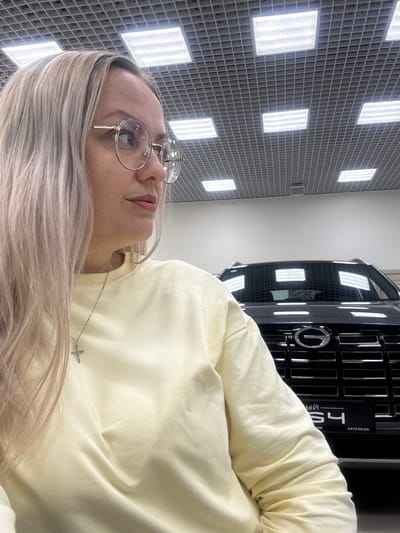 A woman with blonde hair, wearing round glasses and a light yellow sweatshirt, is shown in a selfie-style photograph. A portion of a dark-colored SUV is visible behind her, with its grille and headlights illuminated by overhead fluorescent lighting. The ceiling features a grid of square light panels. The setting appears to be an indoor showroom or garage.