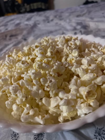 A close-up, high-angle shot captures a generous serving of freshly popped popcorn overflowing from a white bowl. The bowl features a distinct, possibly ruffled or scalloped edge, visible on the right side of the frame. The popcorn is predominantly white, with some kernels displaying a yellowish tint, suggesting butter or oil. The individual pieces of popcorn are fluffy and irregular in shape, indicating a good pop.
The background, largely out of focus, consists of a patterned fabric, likely a blanket or tablecloth. This fabric appears light grey or white with darker grey or black floral or star-like designs scattered across it. On the far left, a blurry dark shape hints at another object or part of the environment, suggesting an indoor setting. The lighting appears warm and diffused, creating soft shadows within the popcorn and highlighting its texture. The overall impression is one of a casual, domestic scene, perhaps indicating a snack time or movie viewing. No people, visible text, or specific time of day are discernible from the image itself, though the artificial lighting hints at evening. The location context provided is Sofronievo, Bulgaria.