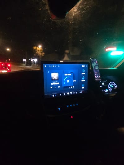 The image displays the interior of a vehicle at night, looking forward through a smudged windshield, situated in Woking, United Kingdom. A central digital infotainment screen, emitting a blue light, dominates the dashboard, showing a music player interface with an album cover, a numerical volume indicator of '17', and a digital clock displaying '10:52'. Below this screen, multiple blue-illuminated control buttons are visible, including a red triangular hazard light button. To the right of the infotainment display, a smartphone in a camouflage-patterned case is vertically mounted. A segment of the vehicle's instrument cluster, featuring a blue-lit dial with an active needle, is also visible on the right side of the frame. Outside the vehicle, the red taillights of another car are visible on the left. The road ahead is illuminated by streetlights, with a yellow-lit tree further in the distance. A prominent bright green traffic light is visible in the upper right portion of the external view. No individuals are depicted within the scene.