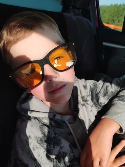 A young boy is sitting inside a vehicle, likely a car, wearing a grey tie-dye hoodie and black Prada sunglasses with orange lenses. Sunlight casts a shadow across the upper portion of his face. The car's interior is visible behind him, including the seat and headrest. In the reflection of the sunglasses, it appears he is traveling through a rural environment. The picture was taken in Diurtiuli, Russia.