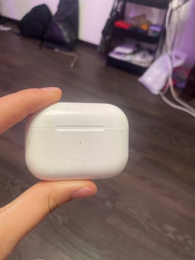 A close-up shot shows a person's thumb and index finger holding a white charging case for earbuds. The case appears slightly dusty. In the background, a blurred image of a room is visible, with furniture and objects that are out of focus. The overall lighting suggests an indoor setting. The provided location of Romanovka, Russia, cannot be visually confirmed from the image.