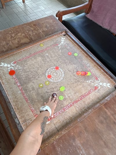 In Ban Hin Chang, Thailand, a carrom board rests on a worn wooden table. The board is marked with red and white decorative elements and has various colored discs scattered across its surface, some with numbers. A hand, adorned with a white watch and a tattoo on the forearm, is poised over a black disc with the number 9. To the right of the table, a dark leather bench with a pink towel draped over its back suggests a casual, outdoor setting. The floor beneath is tiled with a light gray geometric pattern.
