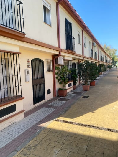 The image shows a row of townhouses in Cancelada, Spain, under a clear blue sky. The facades are painted cream with brown trim along the roofline and window frames. Each townhouse features a dark metal security gate over the entrance door and windows, with some windows also having dark metal bars. Small balconies with black wrought-iron railings are present on the upper floors. Several air conditioning units are mounted on the exterior walls. Potted trees and plants are lined up along the walkway in front of the houses. The walkway is paved with patterned yellow concrete on the right and a combination of red and grey square tiles on the left. A metal grate drain runs across the yellow paved section. The scene appears to be a residential street or courtyard, with no people visible.