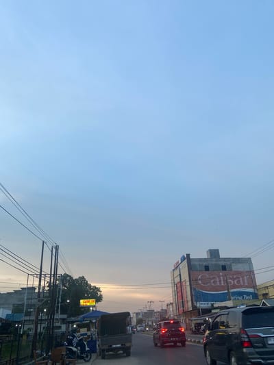 The image captures a street scene in Air Molek II, Indonesia, during twilight. The sky is a pale blue with hints of orange from the setting sun. On the right side of the street, a multi-story building is prominently visible, featuring a large sign that reads "Caisar" and "Spring Bed." Several vehicles, including cars and a truck, are on the road. On the left side, there are more buildings, power lines, and trees. A motorcycle with two riders is parked on the side of the road, along with some chairs. The overall impression is that of a busy urban environment at the end of the day.