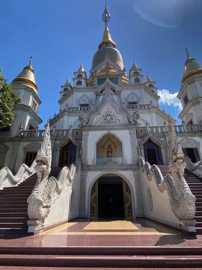 The image captures a low-angle view of a white and gold ornate Buddhist temple structure with intricate detailing. The temple features multiple spires, decorative moldings, and carvings. Two large stone dragons flank the central staircase leading up to the temple entrance. The sky is clear blue with some wispy clouds visible. The building appears to be located in Dĩ An, Vietnam.