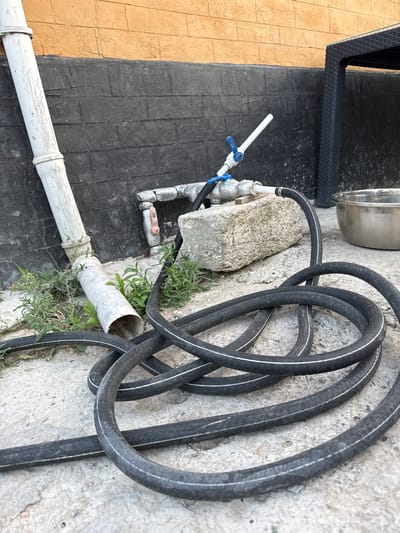 The media file is a top-down shot of an outdoor scene. The main subjects are a coiled black garden hose with white stripes and a white pipe with a blue-handled valve attached. The hose is resting on a textured concrete surface, partially overgrown with small green weeds. Next to the hose is a rough, light-colored rock. The white pipe runs diagonally across the frame, supported by a dark, textured surface that resembles a wall or a large flat object, which transitions to a light brown textured surface. A silver metal bowl or basin is visible in the lower right corner.
The scene appears to be in a backyard or outdoor utility area. There is no clear indication of the time of day or weather, although the lighting suggests it is daytime. The setting in Mastaga, Azerbaijan, is provided as context. There is no activity or event taking place; the objects are static. Notable details include the texture of the surfaces, the coiled nature of the hose, and the details of the plumbing fixture. No visible text is present in the media file.
