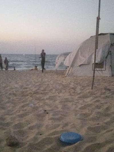 The image depicts a twilight scene on a sandy beach in Al-Mawasi, Palestinian Territories. In the foreground, a blue, disc-shaped object rests on the sand, alongside a darker, irregular object to its left. The midground features several individuals and temporary structures. A person stands facing the ocean, holding a fishing rod. To their left, two additional figures are partially visible near the water's edge, and another figure lies prone on the sand. The ocean shows gentle waves extending towards the horizon, where a faint light source is visible. On the right side of the frame, two light-colored tents are erected on the sand, with one exhibiting visible support poles and ropes. A tall, slender pole stands vertically to the right of the tents. The sky above the ocean displays a gradient from light at the horizon to slightly darker overhead, indicative of dusk or dawn. The overall lighting is low, creating silhouettes for the people and some objects. The sand shows ripples and footprints.