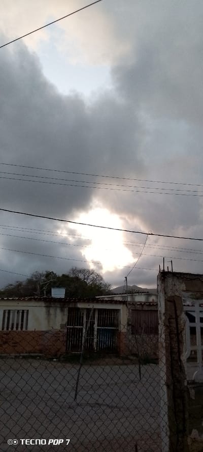 This is a photograph taken from a low angle, looking upwards towards a dramatic sky. The sky is filled with dark, ominous clouds, but there is a bright opening in the center, allowing sunlight to stream through. This creates a striking contrast between light and shadow.
In the foreground, there are several horizontal power lines stretching across the frame. Below the sky, a row of buildings is visible. These appear to be single-story structures with tiled roofs. Some have barred windows and doors, suggesting a residential or possibly a commercial area. A chain-link fence runs along the bottom of the image, obscuring the lower parts of the buildings and the ground in front of them.
The overall mood of the image is somewhat somber due to the dark clouds, but the bright patch of sky offers a glimmer of hope or a focal point. The scene appears to be an outdoor setting, likely a street or a yard, in a populated area. The presence of power lines and buildings suggests a town or city. The time of day is difficult to pinpoint precisely due to the cloudy conditions, but the quality of light suggests it could be late afternoon or early evening, or perhaps before a storm.
Visible text includes "TECNO POP 7" in the bottom left corner, which is likely a camera watermark. The location context provided is Altagracia, Venezuela.