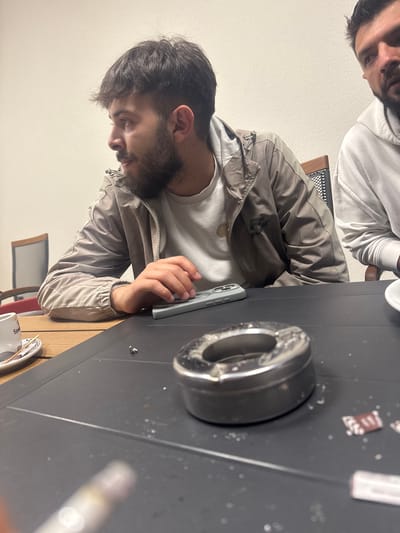 This image, presented sideways but depicting an upright scene, captures two men seated at a dark table in an indoor setting, likely a cafe or dining area.
In the foreground and mid-ground, a man with dark hair and a beard is engrossed in his light blue/mint green smartphone, which he holds in his hands. He wears a light-colored long-sleeved shirt underneath a light brown or tan jacket, which features a dark emblem on the left chest area, possibly reading "FUJI." His gaze is directed downwards at the phone.
Partially visible in the upper left corner of the frame is the face of a second man, showing only his forehead, eyes, and the top of his head. He appears to be wearing a white hoodie.
The dark table in front of the men holds several items. A prominent metallic ashtray is visible, along with scattered small white/grey particles, possibly ash or crumbs. Several small brown paper packets or wrappers, with illegible text, are also on the table. Further to the right, a blurry white object, possibly a lighter or vape pen, is present. A white cup or mug, partially visible on the bottom right, has text on its side that appears to include "Sp...S."
In the background, a plain light-colored wall (white or off-white) provides a simple backdrop. A wooden chair with a dark metal grid backrest is discernible, and another chair with a red backrest is faintly visible further back. The overall atmosphere is casual, and the indoor lighting does not indicate a specific time of day. The provided context indicates the location is Kraishte, Bulgaria.