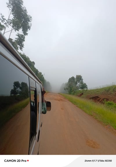 This image captures a perspective from inside a moving vehicle on a rural road, likely in or around Nguroje, Nigeria. The day is overcast and foggy, significantly reducing visibility and creating a diffused light. The road itself is unpaved, appearing to be made of reddish-brown earth, with visible patches of gravel and dirt. To the right of the road, a grassy embankment rises, with some exposed soil and erosion visible. Lush green grass and vegetation line the roadside and the embankment. In the distance, trees and foliage are shrouded in mist, their forms softened by the fog.
The most prominent element on the left is the side of the vehicle the photo is taken from. Its windows are tinted, reflecting the misty environment and some of the trees. A person's arm and hand, wearing a green sleeve, are visible extending from the open window, with the hand resting on the vehicle's frame. The reflection in the window reveals the interior of the vehicle and the person's head and shoulder. The overall atmosphere is one of a quiet, perhaps slightly eerie, journey through a foggy landscape.
