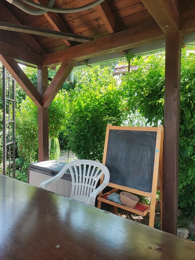 The image depicts a covered outdoor seating area, likely a gazebo or pergola, in the town of Opatovice, Czechia. The structure has a wooden roof and supports, with exposed beams. In the foreground, a dark brown, glossy wooden table is visible, partially obscuring the scene beyond. Behind the table, a white plastic chair with a slatted back is positioned next to a beige storage unit. To the right of the chair is a wooden easel holding a black chalkboard, which appears to have chalk markings. Below the chalkboard, a small red tray holds a wicker basket containing a green cloth and a few other items. The background is dominated by lush green foliage, suggesting a garden or natural setting. The lighting indicates it is daytime, with sunlight filtering through the trees and casting shadows. There are no people visible in the image.