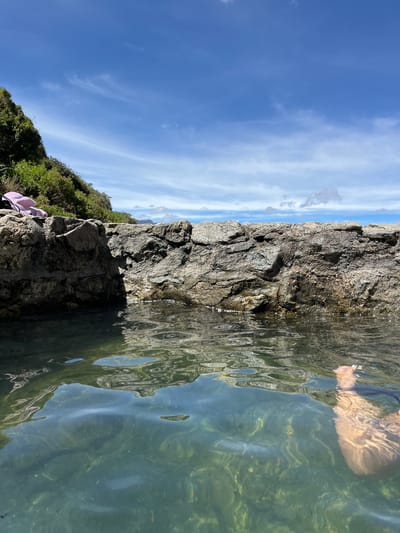A person's legs are submerged in clear, shallow water within a natural rock pool. The water's surface is disturbed by ripples, reflecting the sunlight and the rocky edges of the pool. Beyond the pool, a lush green hillside rises under a bright blue sky dotted with wispy white clouds. A small pile of pink and black clothing rests on the rocks to the left. The scene appears to be an outdoor natural setting, likely in a coastal or mountainous area. This location is identified as being in Mucurubá, Venezuela.