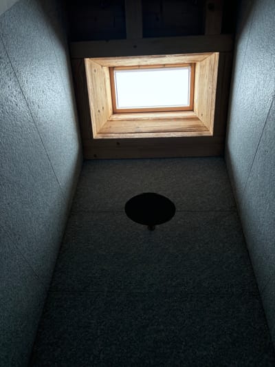 The image shows the interior of a room in Celerina/Schlarigna, Switzerland, located at latitude: 46.514, longitude: 9.859. The room has a skylight with a wooden frame and a view of the sky. There is a black circular object, possibly a light fixture, located on the ceiling, directly below the skylight. The room's walls are made of a dark gray material with a rough texture, and there are no people present. The image is taken from a low angle, looking upward toward the skylight.