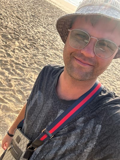 A male subject is depicted in a selfie on a sandy beach, identified as being in the city of Bhagatwadi, India. The subject, an adult male with light stubble and short brown hair, is wearing a patterned grey-brown bucket hat with "CHRISTIAN DIOR PARIS" partially visible, silver-framed sunglasses reflecting the environment, and a dark grey t-shirt with a subtle repeating pattern. A beige shoulder bag, featuring a repeating logo pattern resembling the Gucci GG monogram, is slung across his body with a navy and red striped strap. A dark wristband is visible on his left wrist. He is smiling and looking directly at the camera. The background consists of light-colored sand with ripples and subtle footprints. The ocean is visible in the upper right portion of the frame. Lighting suggests daytime conditions.