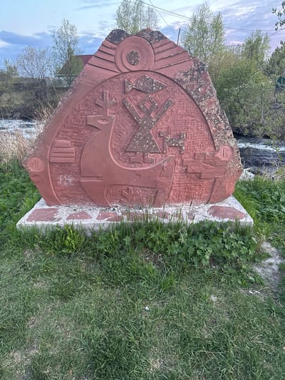 The image shows a large, red stone monument covered with etched designs. The monument is placed on a base made of lighter-colored, possibly concrete, stones. The monument and its base are surrounded by grass and some other greenery. A blurred background reveals trees, a waterfall, and a glimpse of the sky. The monument appears to be located outdoors, likely in a park or other natural setting near the waterfall. The location is Luven'ga, Russia.