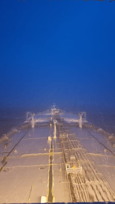 An aerial, high-angle, slightly blurry, vertical shot depicts the deck of a large tanker ship at dusk. The sky is a deep, uniform blue with some scattered white particles that could be rain or spray. The ship's deck is metallic and reflects the dim ambient light, creating a somewhat glossy appearance. Numerous pipes, organized in parallel rows, dominate the right side of the deck, running towards the bow. There are also several structures resembling ventilation shafts or equipment housings visible on the deck, particularly towards the center and front of the ship. The overall impression is one of industrial infrastructure at sea under a twilight sky. The image does not contain any information about a specific city in Sri Lanka.