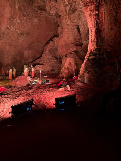 The image depicts an indoor scene within a cave illuminated by red lighting. In the background, several individuals dressed in orange garments and one person in a red jacket are standing or observing. Some figures are positioned near a large rock. In the middle ground, a person is lying face down on the ground, with a red rope attached to their body. Several pieces of equipment, including what appear to be lights and a control panel, are placed on the red, gravelly floor. The environment is characterized by rough, textured cave walls and a floor covered in red gravel. The scene appears to be a staged or artistic setup, possibly for a performance or photoshoot, given the lighting and props. The specific location in Guttapadu, India, is not visually evident from the provided image.