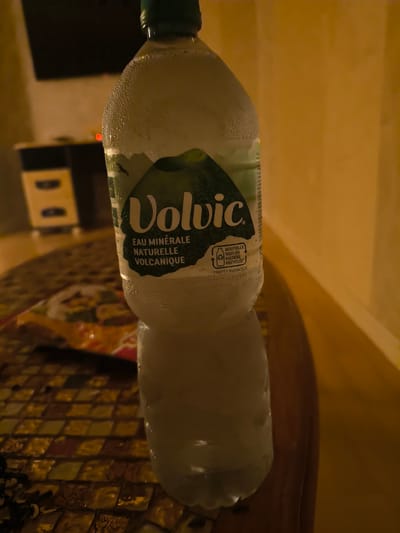 A close-up, waist-level shot shows a condensation-covered bottle of Volvic mineral water positioned on a mosaic-tiled surface in Le Mans, France. The bottle's label features the "Volvic" logo in white text against a dark green background, with additional text indicating "EAU MINÉRALE NATURELLE VOLCANIQUE" and a recycling symbol with the text "BOUTEILLE 100% EN MATIÈRE RECYCLÉE". The background is dimly lit, with a blurred view of furniture and a wall.