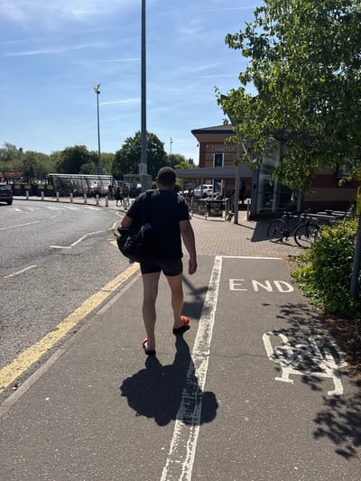 A man walks on a marked bike path in Colchester, United Kingdom. The man, facing away from the camera, wears a black shirt, black shorts, and red running shoes. The bike path has a white "END" marking, and a bicycle symbol. Behind the man is a building labelled "Charter." A bicycle rack is nearby. The sky is blue with wispy clouds. A lamppost is visible.