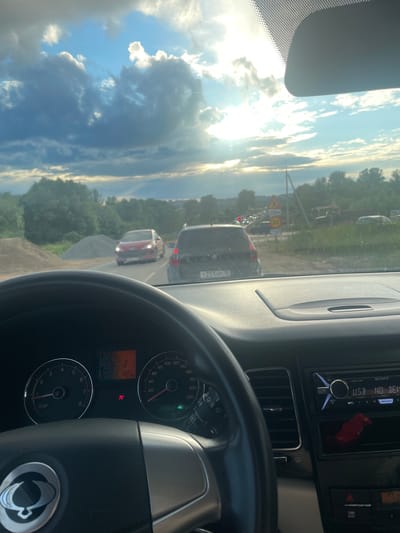 This media file captures a view from the driver's seat of a car on a road in Gavrilovka, Russia. The sun is shining brightly, casting long shadows and illuminating the sky with dramatic clouds. The time of day appears to be late afternoon or early evening, given the angle of the sun and the lighting conditions.

The scene depicts a traffic jam. Several cars are visible ahead, including a dark SUV and a red car. Road signs indicate traffic regulations, with one sign showing "250m" and another displaying a speed limit of "30" and the word "СТОП" (STOP). The car's dashboard is visible, featuring a speedometer reading around 20 km/h and a temperature gauge showing 19 degrees Celsius. The car stereo, a Sony model, is also visible, displaying "USB NO DEV."

The overall impression is one of a typical drive on a Russian road, with the challenges of traffic and the beauty of the natural surroundings. The presence of the traffic jam suggests a possible commuter route or a congested area. The weather appears to be fair, with a mix of sun and clouds. The photo is taken from a first-person perspective, immersing the viewer in the experience of being in the car. The license plate of a car ahead is partially visible, reading "T251HP 10."