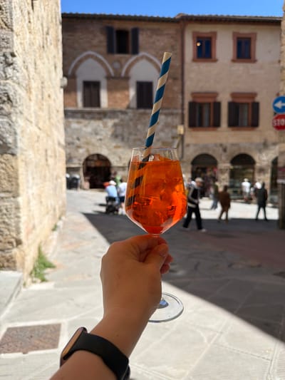 The image, when viewed upright, presents a first-person perspective of a hand holding a refreshing orange-colored drink. The drink, likely an Aperol Spritz, is served in a stemless wine glass filled with ice cubes and features a blue and white striped paper straw. The hand, with manicured fingernails, has a black smartwatch on its wrist.

The scene unfolds on a sunny day in what appears to be a historic European town, specifically San Gimignano, Italy, as indicated by the location context. In the background, traditional stone buildings with multiple windows line a narrow street. The windows feature dark brown wooden shutters, some open and some closed. Arched doorways or passages are also visible within the building facades.

Blurred figures of people can be seen walking further down the street, adding to the bustling atmosphere of the town. Strong sunlight casts distinct shadows on the stone ground, highlighting the clear and bright weather conditions. Visible details include a blue circular traffic sign with a white downward arrow and a partially visible red octagonal "STOP" sign mounted on one of the buildings. A clear blue sky is visible at the top left of the frame. The image conveys a sense of relaxation and enjoyment in a picturesque, historical setting.