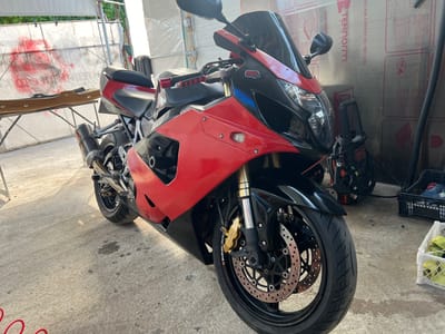 The image depicts a red and black Suzuki motorcycle parked on a concrete floor in what appears to be an indoor or covered space. The motorcycle has aftermarket exhaust and a black windscreen. The surroundings include a table with some object on it in the background, plastic sheeting on walls. A black and decker pressure washer sits behind the bike. The image does not provide information about the city of Karnalovo, Bulgaria. There are no people visible in the image.