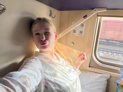 A woman with blonde hair, wearing a floral-patterned blouse, is pictured inside a train carriage, specifically in the city of Krasnyy Sulin, Russia. She is positioned with her head down, and is winking at the camera. A window is visible to the left, with a train car visible. A seat is below, with a fold out tray. QR codes and signs are to the right.