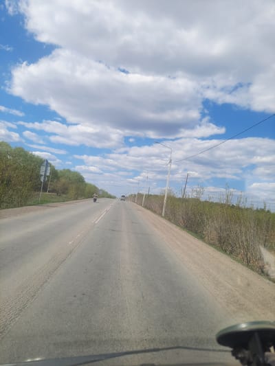 The image depicts a straight asphalt road stretching into the distance, likely taken from within a vehicle. A motorcyclist is visible in the distance, and a vehicle further ahead. Power poles line the left side of the road, with trees visible beyond them. The sky is blue with white, puffy clouds. The right side of the road has a more overgrown, grassy shoulder. The location is in Научный Городок, Russia.