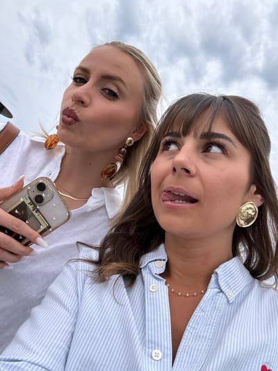 Two young women are posing for a selfie outdoors. The woman on the left has blonde hair pulled back, fair skin, and is making a pouting expression. She wears large amber-colored earrings, a gold necklace, and a white button-down shirt. She holds a smartphone with a clear case containing a vintage-style Polaroid photo decorated with small hearts. The woman on the right has brown hair with bangs, brown eyes, and fair skin. She is sticking out her tongue slightly and glancing to the side. She wears large gold button earrings and a light blue and white striped button-down shirt with a delicate pearl necklace. The sky is overcast and cloudy. The location is assumed to be somewhere in the city of Vallauris, France, based on the prompt.
