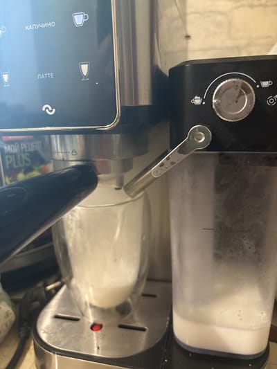 A close-up, eye-level shot shows a coffee machine with a frothing pitcher on the right and a double-walled glass with frothed milk on the left. The coffee machine's digital display is partially visible on the top left, showing options like "КАПУЧИНО" (Cappuccino) and "ЛАТТЕ" (Latte). A dial and buttons are present on the black part of the machine on the right. The scene appears to be indoors, likely in a kitchen, given the presence of the appliance and the background wall. The lighting is ambient and focused on the machine. The image quality is moderate, with some graininess and reflections. The location of Zubov, Russia, is not discernible from the visual elements present in the image.
