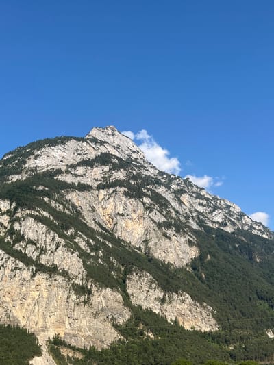 The image depicts a large mountain located in the city of Schattdorf, Switzerland, under a clear, bright blue sky. The mountain's facade is predominantly composed of various shades of light grey, white, and pale brown rock, exhibiting distinct horizontal and diagonal geological striations and rugged textures. Interspersed and growing upon these rocky surfaces are numerous sections of dense, dark green coniferous trees, particularly prevalent on the lower and mid-slopes. The mountain culminates in a jagged, lighter-toned peak toward the upper center of the frame. A few small, scattered white cumulus clouds are visible in the blue sky, primarily near the mountain's peak and along its upper right slope. Faint, dark horizontal lines, consistent with power or communication lines, traverse the lower right section of the mountain's forested area. No human subjects, animals, or other significant man-made structures are present in the scene.