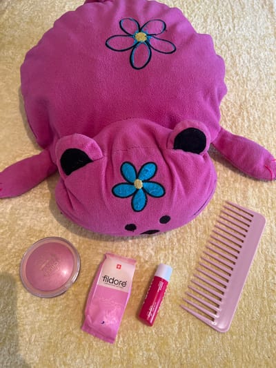 The image shows a pink stuffed animal bear with blue embroidered flowers, a pink comb, a red and white lip balm, a pink packet labeled "fildore LABORATORIE NATUREL," and a pink blush compact on a yellow, textured surface. The items are arranged randomly, suggesting a personal, informal setting. The location, Kvemo Arkevani, Georgia, is not discernible from the image content.
