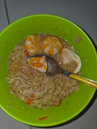 A top-down, close-up shot shows a green plastic bowl containing noodles, broth, egg slices, and orange and white spiral-patterned fish balls. A metal spoon with a yellow handle rests within the bowl. The bowl sits on a light grey tiled surface. The scene appears to be indoors, illuminated by artificial light. The image was taken in Pintomakmur, Indonesia.