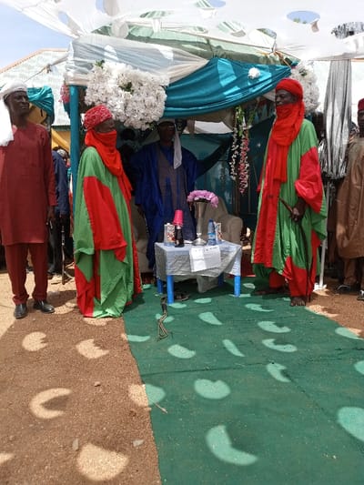 The scene takes place in Zum, Nigeria, at the coordinates 10.116 latitude and 13.047 longitude. There are several men gathered under a canopy tent decorated with white flowers and hanging greenery. The man on the far left is wearing a maroon robe and a white cap. He is standing on a dirt path. The man in the center is wearing a blue robe and a white cap. He is seated behind a table on a green carpet. The table is covered with a white tablecloth and has several items on it including a red cup, bottles of beverages, and a silver vase with pink flowers. The man to the right is wearing a red and green robe and a white cap. He is standing to the right of the table on the green carpet. Other men are standing behind the three men, mostly obscured by the tent. The canopy is white with a blue lining. Sunlight is shining, creating circular patterns on the green carpet.
