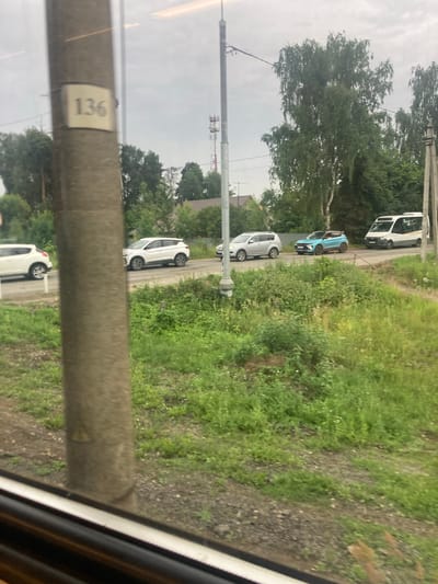 A street scene is captured from a window, showing a number of cars parked and moving along a road. In the foreground, a thick, textured concrete pole dominates the left side of the frame, with a small, light-colored plaque bearing the number "136" attached to it. The pole is out of focus, suggesting the photo was taken from a moving vehicle.
Beyond the pole, a paved road runs horizontally across the middle ground. Several cars are visible on the road. On the left, a white SUV is parked facing right. Further down the road, a white crossover SUV is in motion, also facing right. Next to it, a bright blue, sporty-looking car is driving. On the far right, a white van with yellow trim is driving away from the camera.
The environment is lush and green, with abundant vegetation lining the road. Tall trees with full foliage form a backdrop on either side of the road. In the distance, a cell tower with red and white sections can be seen rising above the trees, and a few residential buildings with peaked roofs are scattered among the greenery. The sky is overcast and appears cloudy, suggesting a temperate climate. The presence of the number "136" on the pole and the context of rural Russia suggest a location in the village of Sokolovo, Russia, possibly a section or route number.