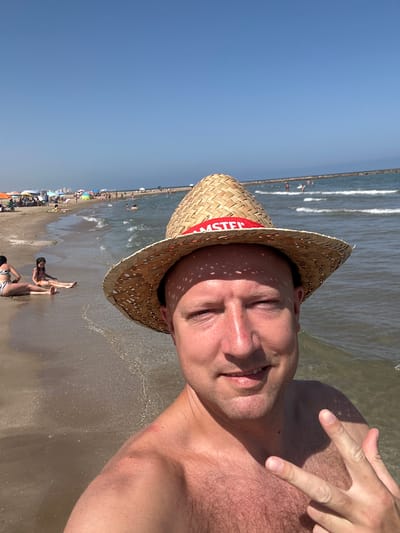 A man wearing a straw hat with a red and white band that says "MASTER" is taking a selfie on a beach. The hat casts a shadow on his face, and his eyes are closed. He is holding up two fingers in a peace sign. The man is shirtless, and the bottom half of the image shows his chest and stomach, which are covered in hair. He is standing in shallow water, with waves breaking on the shore behind him. The beach is crowded with people, many of whom are under colorful umbrellas. The sky is clear and blue, suggesting it is a sunny day. The overall mood of the image is relaxed and joyful, consistent with a day at the beach. The location is Meliana, Spain, implying a Mediterranean setting.