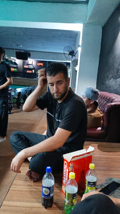 In a dimly lit indoor space, possibly a recreation room, a man with short dark hair and a beard sits cross-legged on a wooden floor, wearing a black t-shirt and dark pants. He is scratching his head, looking downwards with a pensive expression. In front of him are three plastic bottles of drinks, one dark brown, and two lighter colored with green labels, along with a red cardboard box of "Choco-Pie". To his right, a person wearing a light-colored hat is seated on a brown leather sofa, with their back to the camera. In the background, a pool table is visible, along with a digital display showing "01:20:04". A wall fan is mounted on the ceiling. The overall ambiance suggests a casual gathering or downtime, possibly in the city of PistaMador, Tajikistan.