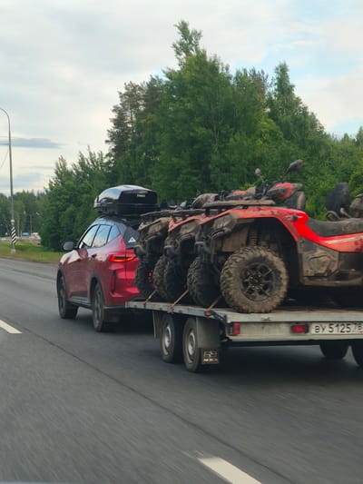 A red SUV with a black Thule roof box is towing a trailer loaded with four red and black ATVs on a highway. The ATVs are covered in mud. The license plate on the trailer is visible and reads "BY 5125 78". The scene is set in a wooded area, with a cloudy sky overhead. The image appears to have been taken from another moving vehicle, as indicated by the motion blur. The location is specified as Okhtinskoye Razdol'ye, Russia.