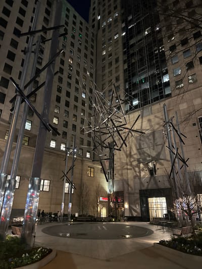An abstract sculpture of tangled silver rods and wires hangs in a courtyard. Tall buildings surround the courtyard, reflecting light from numerous windows. The ground is paved with light-colored stone. In the distance, there are signs and some greenery. The scene is at night, with the sky visible as a dark hue. The overall impression is urban and modern.