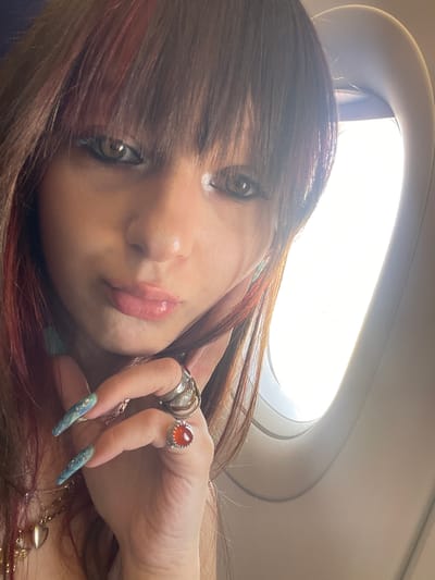 This is a close-up, slightly blurred, selfie-style photograph of a young woman with long, dark brown hair with reddish highlights, posing next to an airplane window. The woman is looking towards the camera with her head tilted slightly. She is wearing makeup, including eyeliner and mascara, and has glossy pink lipstick on her lips. She has long, pointed fingernails painted a light blue with glitter. Her right hand is raised, with several rings on her fingers, including one with a large orange stone and another with multiple bands. She is wearing a necklace with a heart-shaped pendant. The background shows the bright interior of an airplane cabin, with the curved edge of the window frame visible on the right side of the image. The lighting suggests it is daytime, with bright light coming from outside the window. The image does not contain any visual elements that would indicate a specific location in the city of Yagel, Israel.