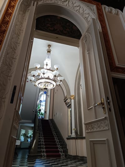 The image shows a wide-angle view of an ornate interior staircase and hallway. A grand, multi-tiered crystal chandelier hangs from a high, arched ceiling. The staircase, covered with a red carpet featuring a decorative border, ascends to the right, with wrought-iron railings. The floor in the foreground is a black and white checkerboard pattern. An open doorway with carved details is visible on the right side of the frame. To the left, an archway frames the view further down the hallway, where plants are visible in the distance and a sign with Cyrillic text that reads "ВХОД НЕТ" (No Entry) is posted. The architecture suggests a historic building, possibly a public institution or a stately home. The image is taken from a low angle, emphasizing the height and grandeur of the space.