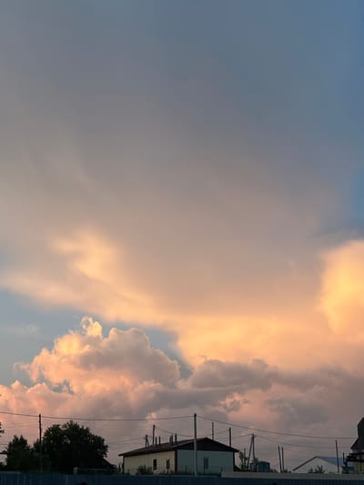 The image shows a twilight sky over the city of Ludorvay, Russia, with large, fluffy clouds illuminated by the setting sun, casting a warm, orange glow. The sky transitions from a soft blue at the top to a hazy, rosy hue towards the horizon. Silhouetted against the sky are a few simple, low-rise buildings with dark roofs, suggestive of a rural or suburban setting. Electrical wires stretch across the scene, supported by utility poles. To the left, a dense cluster of dark green trees stands out. A blue fence runs along the bottom of the frame, partially obscuring the base of the buildings and trees. The overall impression is one of a peaceful evening scene in a quiet locality.