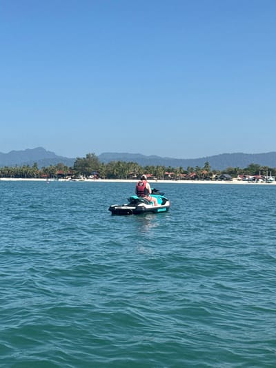 The image depicts a bright, sunny day at a tropical beach location. The perspective appears to be from slightly above or from the shore looking out towards the water.

In the foreground and midground, a person is on a jet ski in the blue-green ocean water. The jet ski is predominantly black and white with teal accents. The brand name "SEA-DOO" is visible on the side of the jet ski. The person on the jet ski is wearing a red life vest and appears to be looking towards the left side of the frame (away from the shoreline). Their posture suggests they are either preparing to ride or have just stopped. The water around the jet ski shows small ripples and waves, indicating a slight breeze or gentle movement.

The background features a long, sandy beach lined with numerous palm trees and other tropical vegetation. Behind the beach and foliage, there are various buildings, possibly resorts or houses, partially obscured by the trees. Further in the distance, a range of hazy blue mountains stretches across the horizon, adding depth to the landscape.

The sky occupies the upper portion of the image and is a clear, bright blue, indicating good weather. The overall impression is one of a leisure activity in a beautiful, warm, and sunny vacation destination.