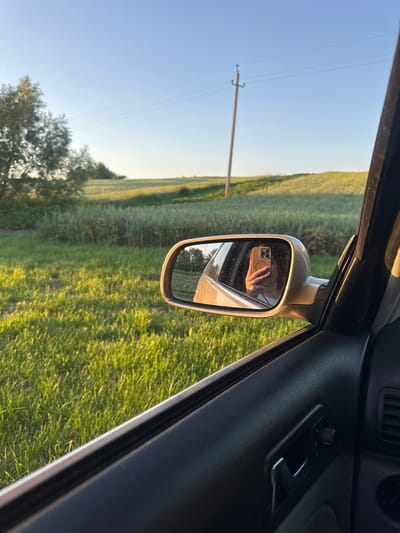A person's hand, adorned with a ring, holds a smartphone in the side-view mirror of a car. The phone's case is a light brown color, and the screen displays a selfie. The mirror reflects the passenger-side interior of the car, including a black door panel, and the lush green field and distant treeline visible through the car window. A utility pole and power lines are visible in the background, against a clear blue sky. The scene appears to be set in a rural environment, possibly on the outskirts of the city of Tovstoluh, Ukraine. The golden hour lighting suggests it is late afternoon or early evening.