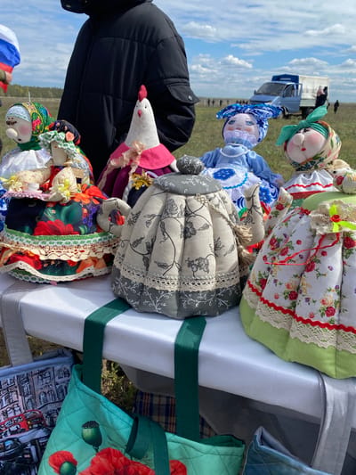 The image depicts a collection of handmade fabric dolls and other fabric goods displayed on a white table outdoors. The scene appears to be at a market or fair. The dolls are dressed in colorful and patterned fabrics with lace and other embellishments. A stuffed goose with a pink collar is also among the items. There are tote bags with various patterns also on the table. In the background, there is a partial view of a black jacket and people walking in the distance and a blue and white vehicle. The sky is visible in the background and appears to be partly cloudy. A Russian flag is visible. The location is in Kumakskoe, Russia.