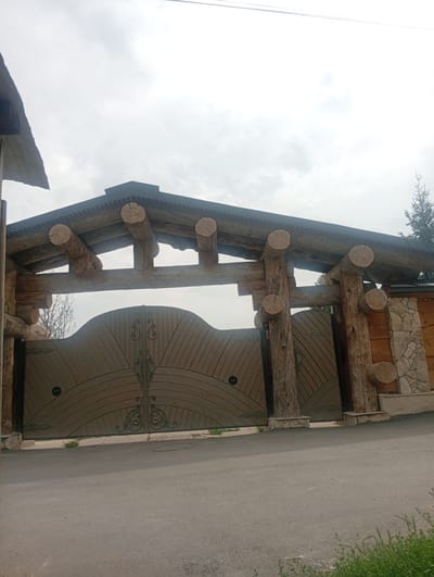A wide shot captures a large, rustic wooden gatehouse with a pitched roof supported by rough-hewn logs in Tash-Moinok, Kyrgyzstan. The gate itself is made of solid, arched wooden panels with decorative metal scrollwork at the center. The log structure is substantial, with horizontal beams and vertical posts that are unvarnished and show signs of weathering. Some of the logs have been cut to display their circular cross-sections. The gatehouse is flanked on the right by a wall constructed of stone and wood. In the background, a conifer tree is visible against a cloudy sky. The foreground consists of a paved road leading up to the gate, with a patch of green grass and foliage on the right side of the road. No people are present in the image. The overall impression is one of a sturdy, naturalistic entrance to a property.